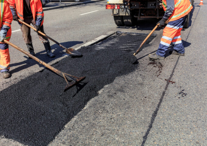 The working team of road builders distributes fresh asphalt on the road section and levels it for repair.